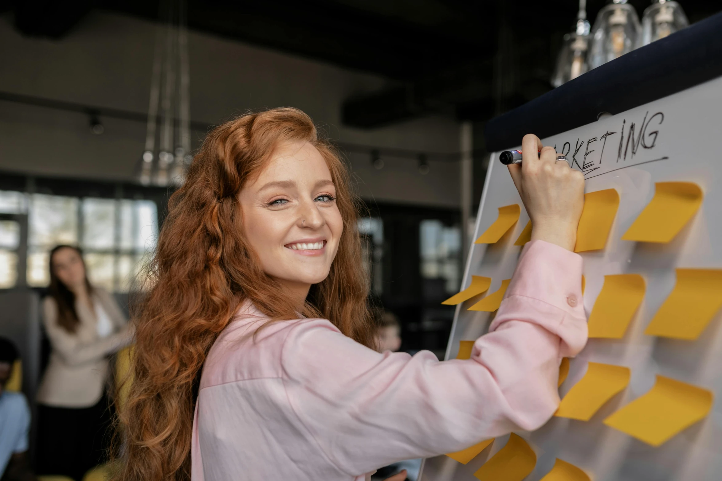 a woman with curly red hair and a smile is writing on a piece of yellow tape