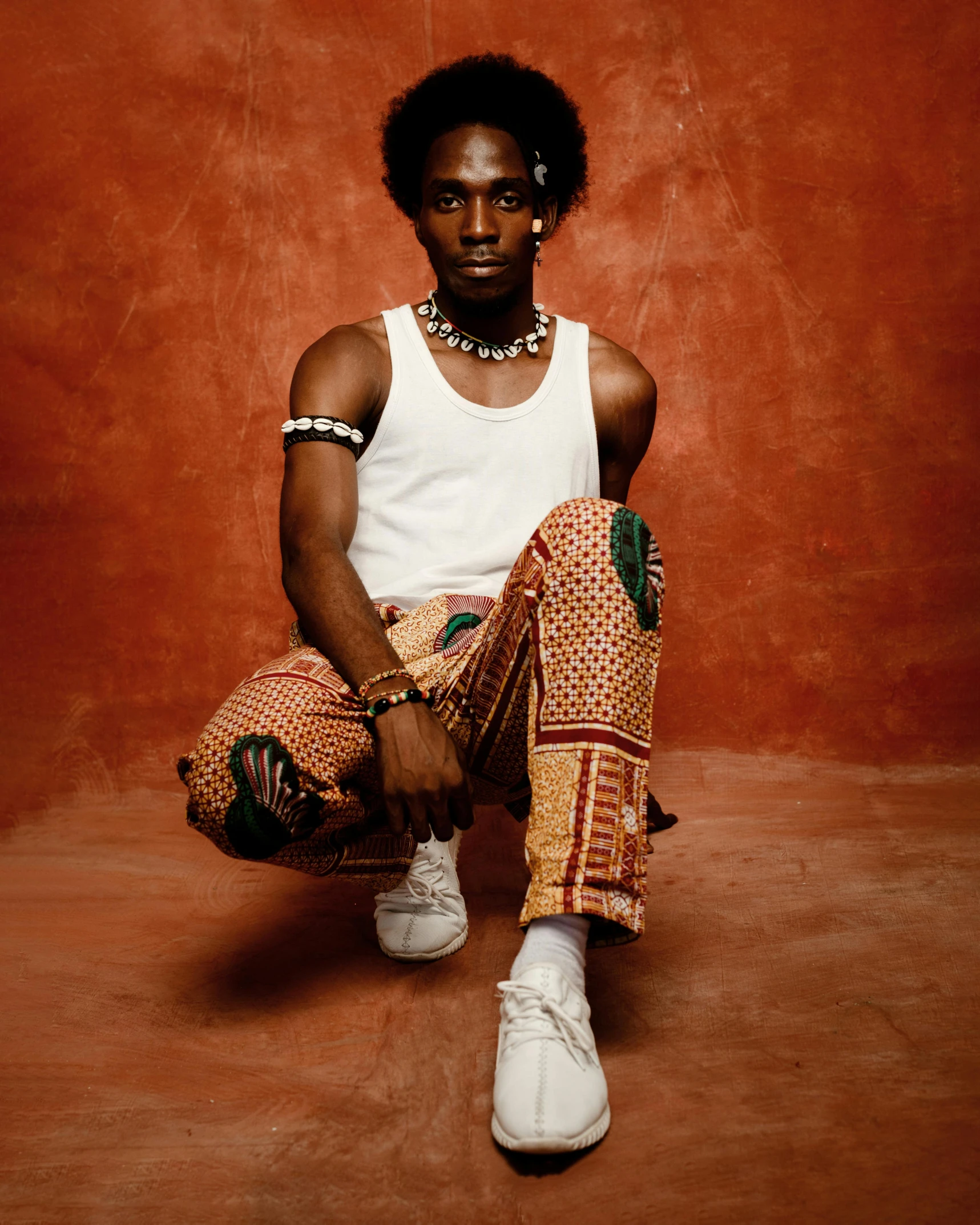 a man wearing a white tank top and patterned pants sitting in a studio