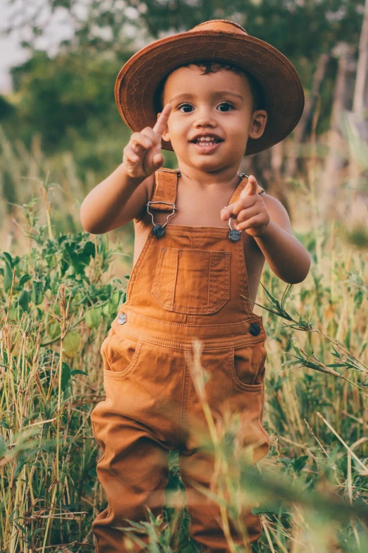 small boy wearing overalls and a hat standing in tall grass