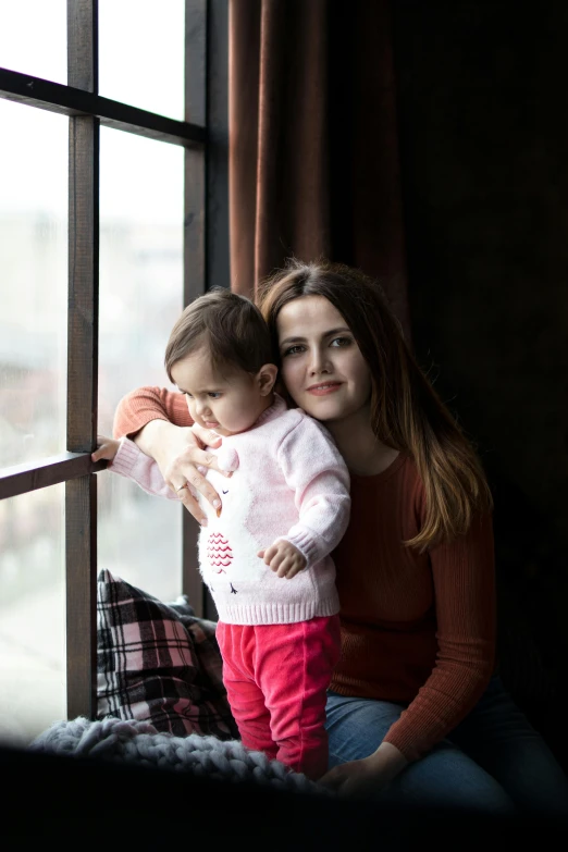 a young woman holding her baby girl while sitting in front of a window