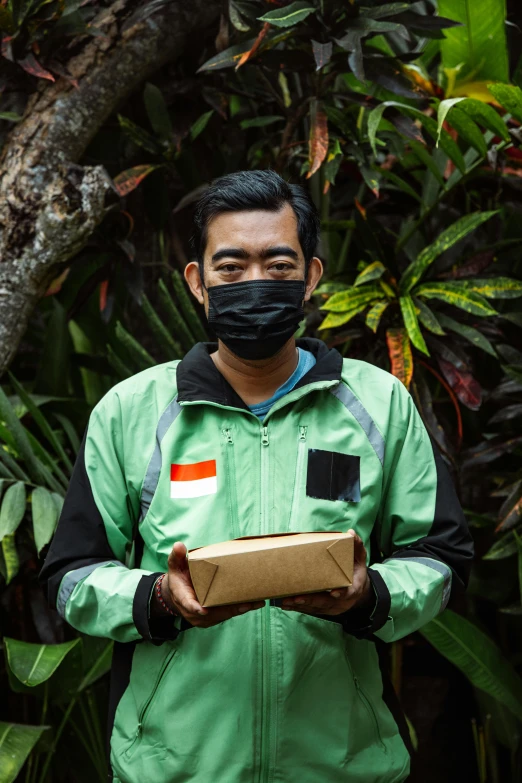 a man in a face mask and green jacket holding a cardboard box