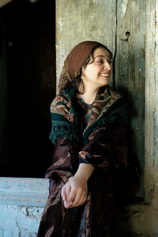 a smiling woman sits in the doorway while wearing a brown hat and coat