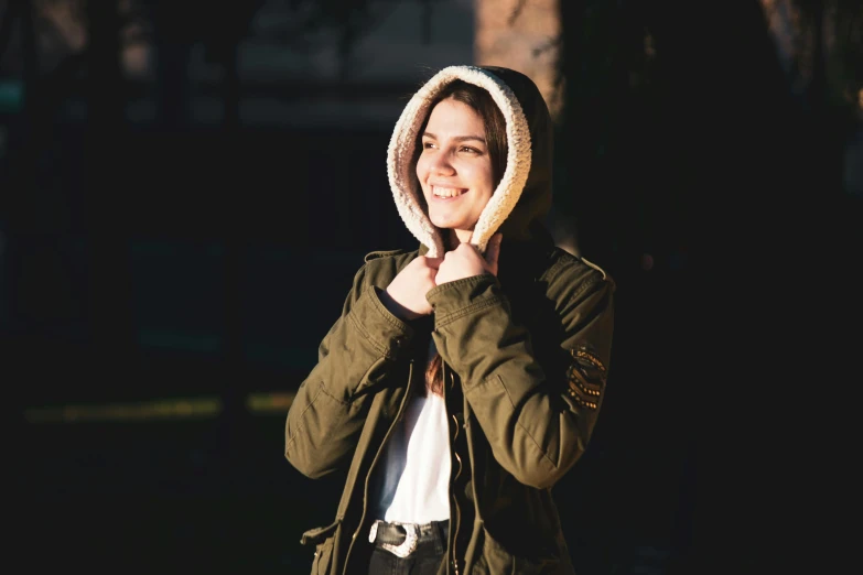 a woman standing in front of a dark forest, wearing a green jacket and hood