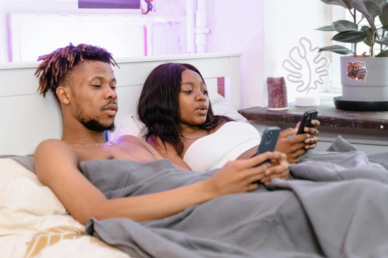 two young adults are using cell phones in bed