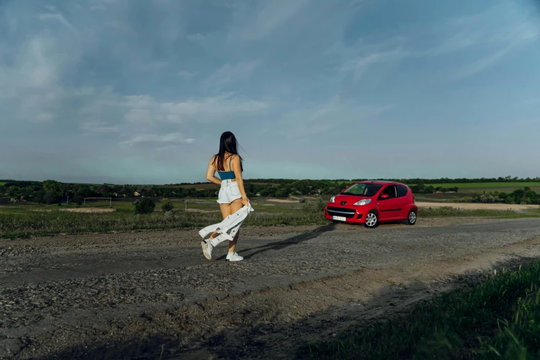 a girl in white dress walking on dirt road next to red car