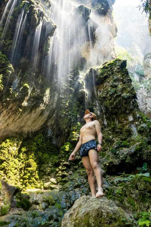 a man in swim trunks standing near a waterfall