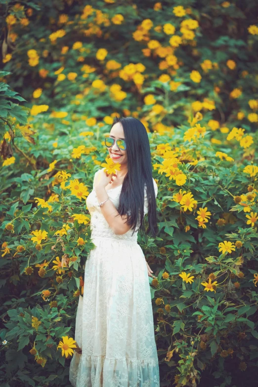 a woman with long dark hair and white sunglasses stands among yellow flowers