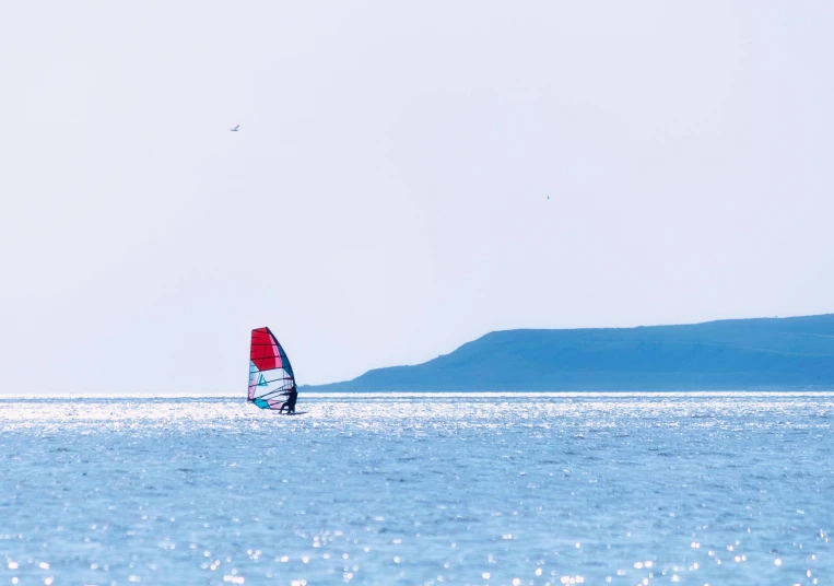 a sail boat sailing across the water in front of a small hill