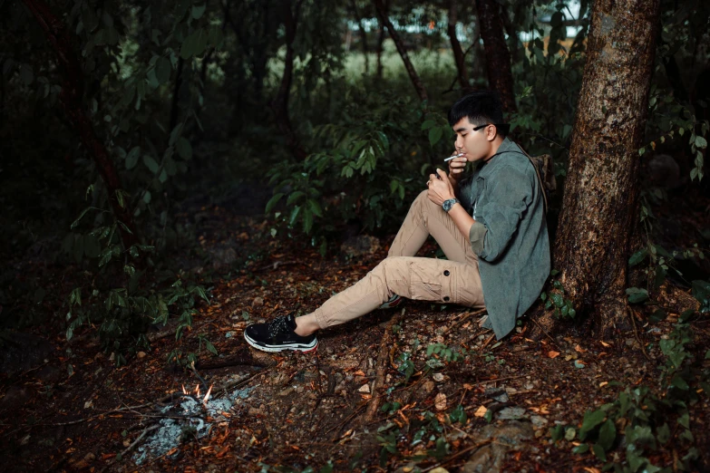 man sitting on the ground smoking cigarette next to trees