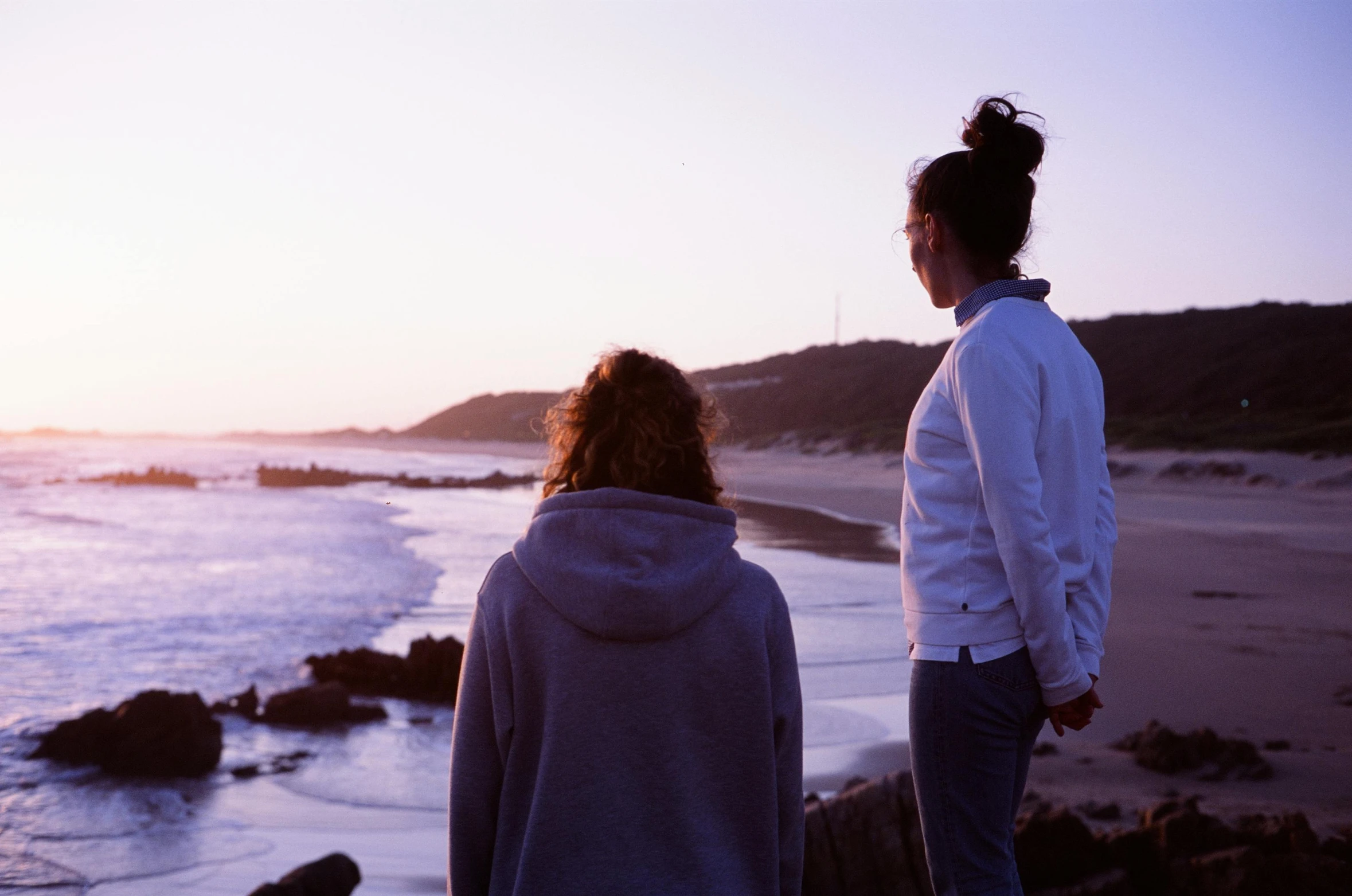 two women stand at the edge of a beach watching the sunset