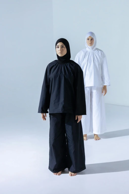 two women in a white room, one in black with a black head scarf and one wearing a black coat