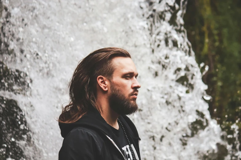 man with long hair standing in front of waterfall