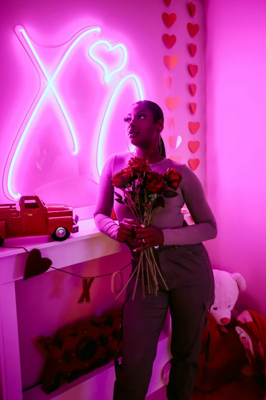 a woman in an all - grey outfit stands next to a pink light and displays some flowers