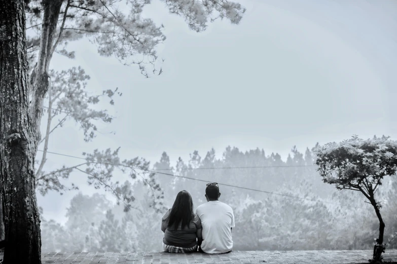 a couple is sitting on the ground in front of a tree looking at the woods