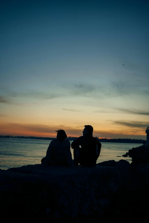 two people sitting on a rock by the ocean at sunset