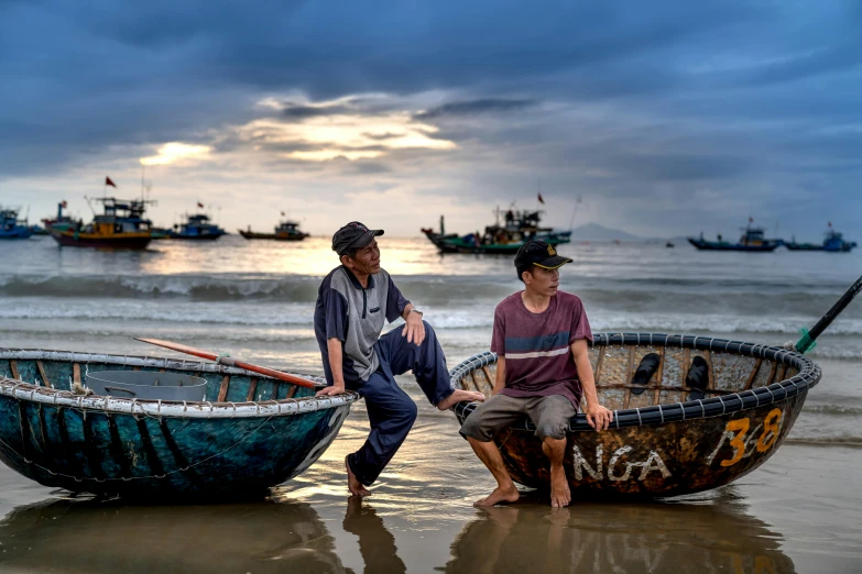 two people are sitting on the beach near two boats