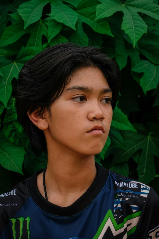 a boy with black hair stands in front of some green plants