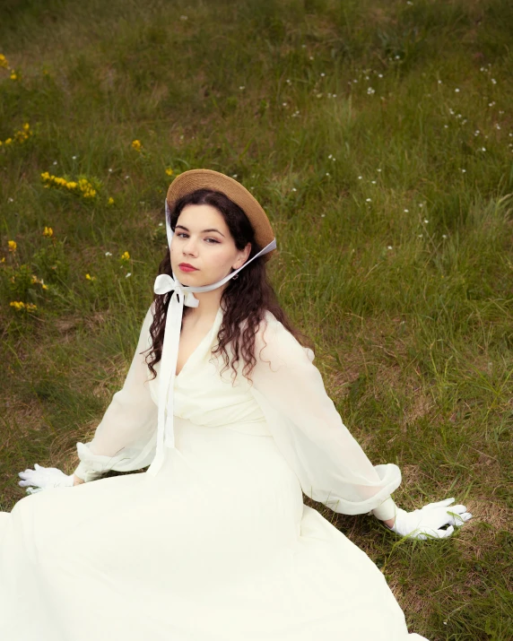 a woman wearing a dress and hat, seated on the grass with her hands in her hair