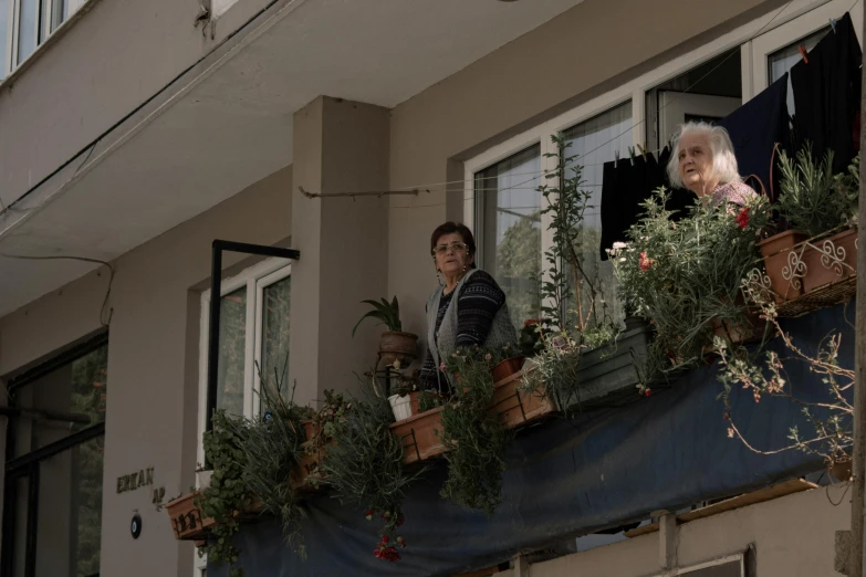 an elderly woman on a balcony with flowers in planters