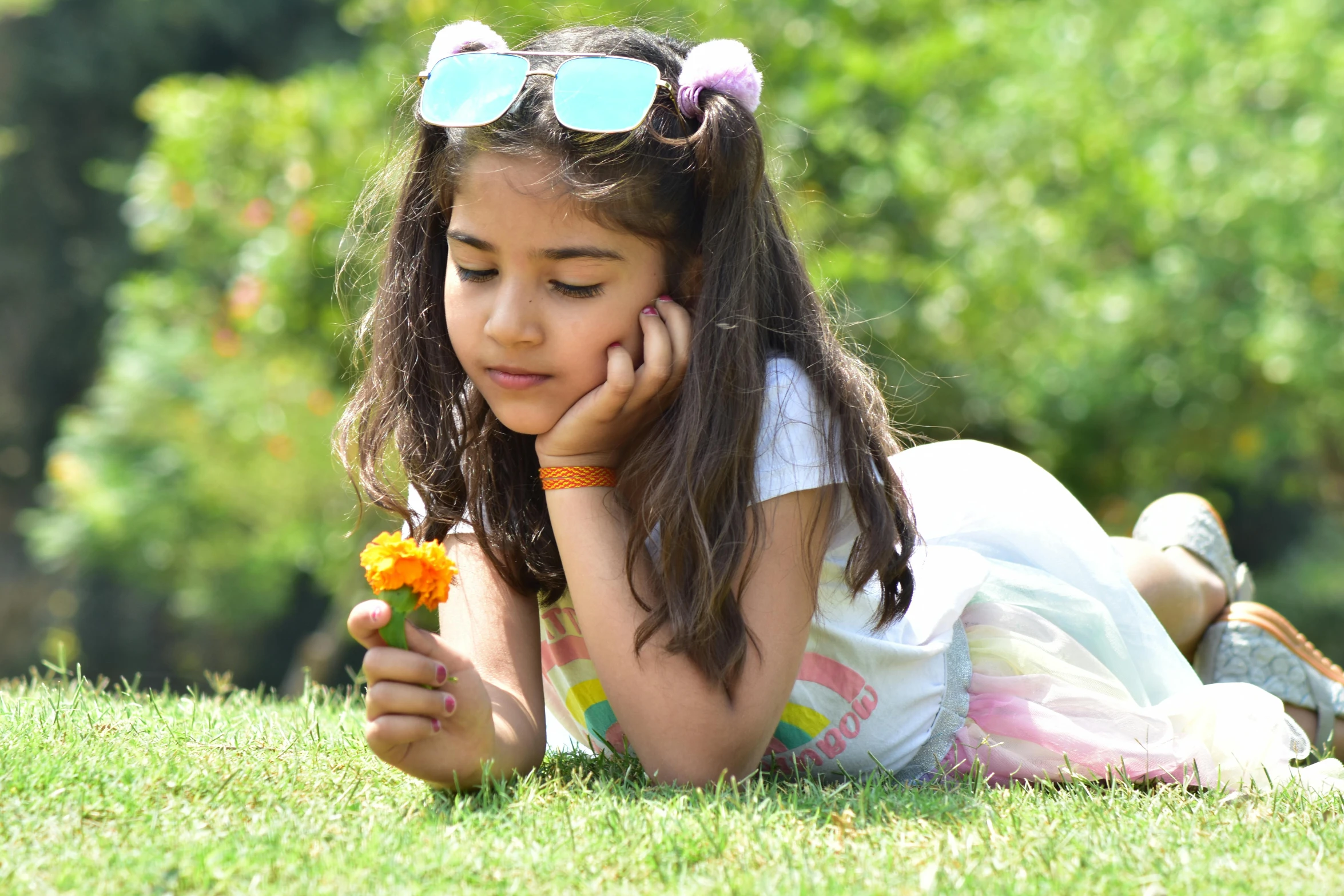 a small girl in sunglasses is laying on the grass looking at a flower