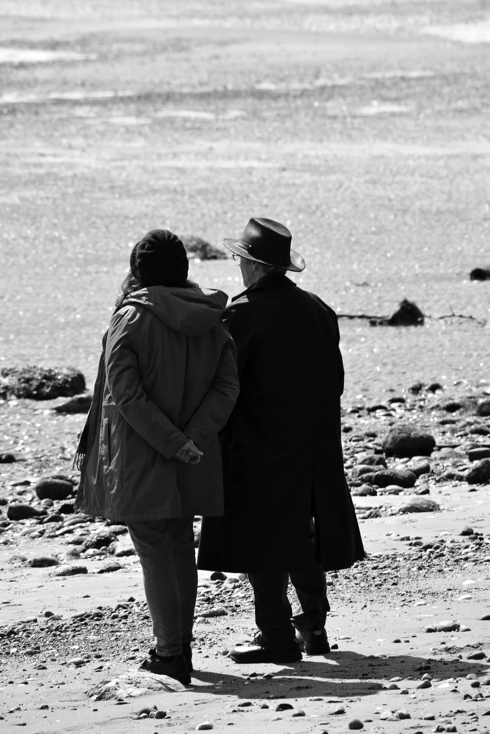 two older people stand on a beach looking at the ocean