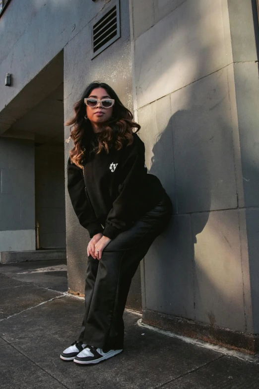 a woman sitting against a wall wearing sunglasses and jacket