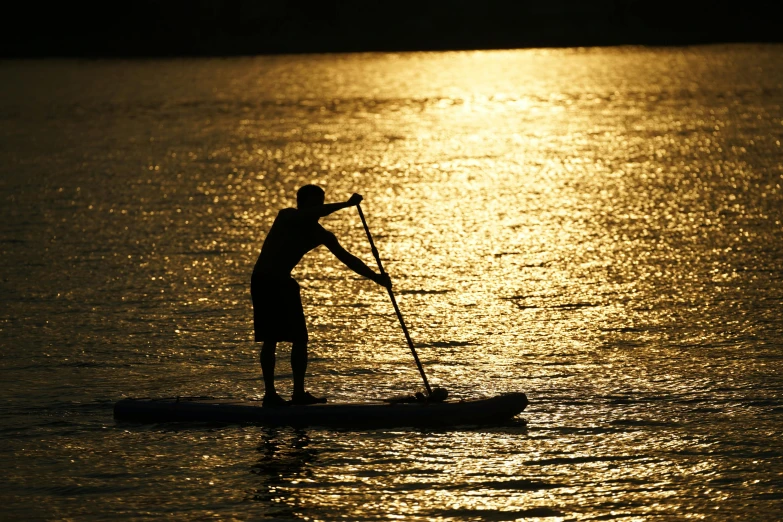 man paddle boarding on calm body of water with bright sun shining behind