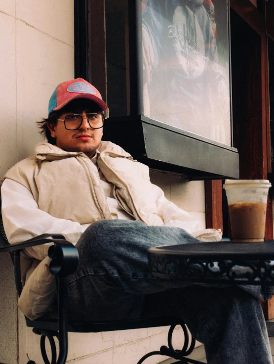 a young man is sitting in a chair with a cap and drinking coffee
