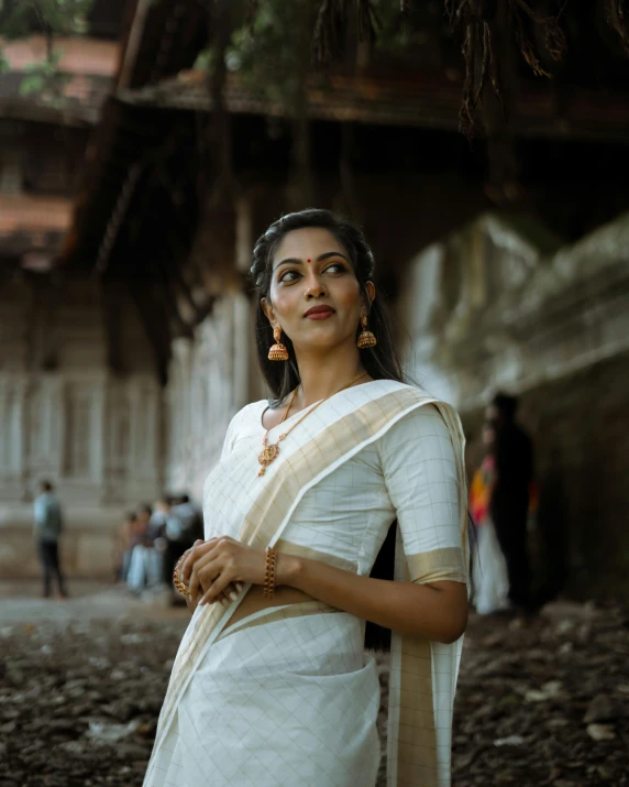 a woman in white and gold sari, stands in front of a stone structure