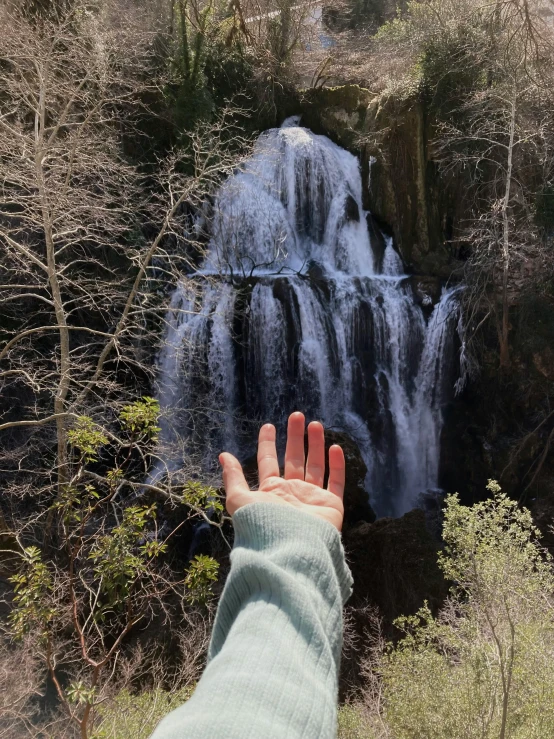 person's hand reaches out for waterfall in the distance