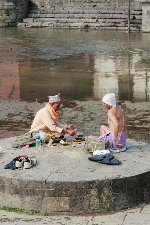 two men sitting in front of a river on a concrete surface