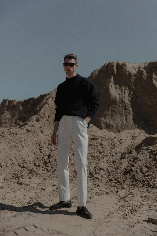 a man wearing a black sweater and white pants standing on sand