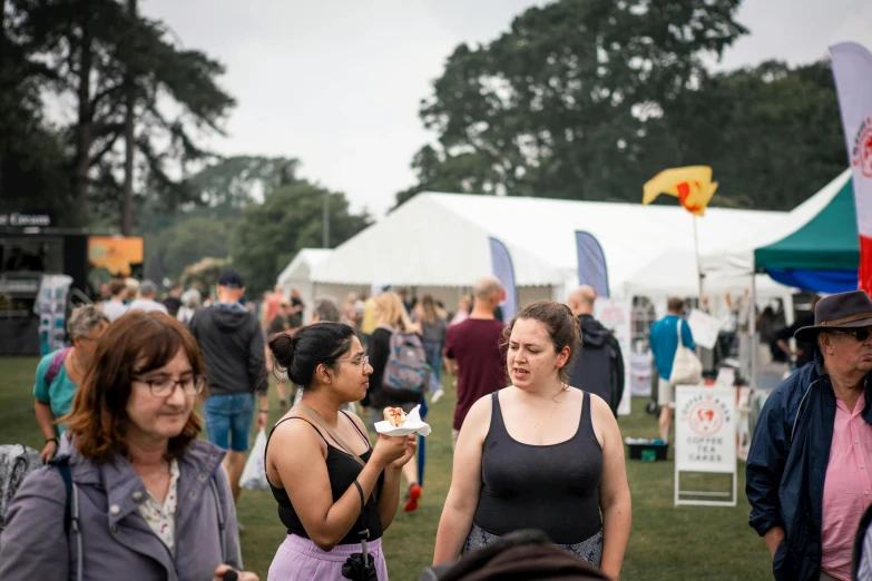 several women at an outdoor festival, one holding food and the other looking into the camera