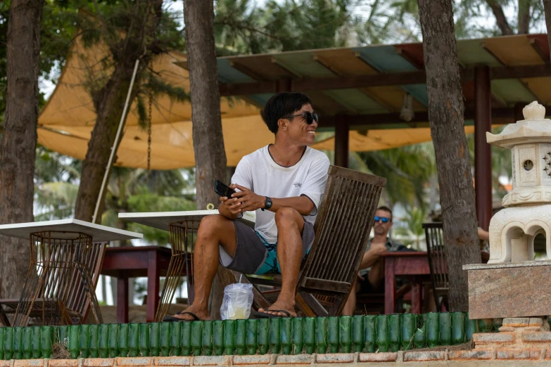 a man sitting on a chair in the shade with a cellphone