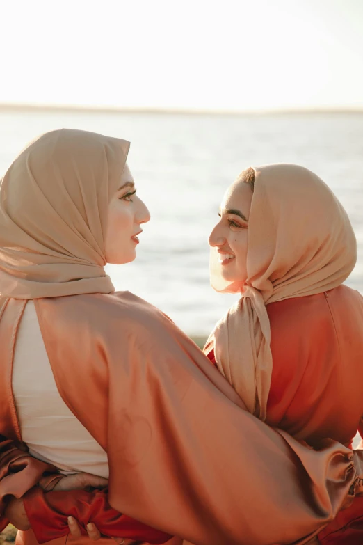 two women in headscarves sit on the shore looking out at the ocean