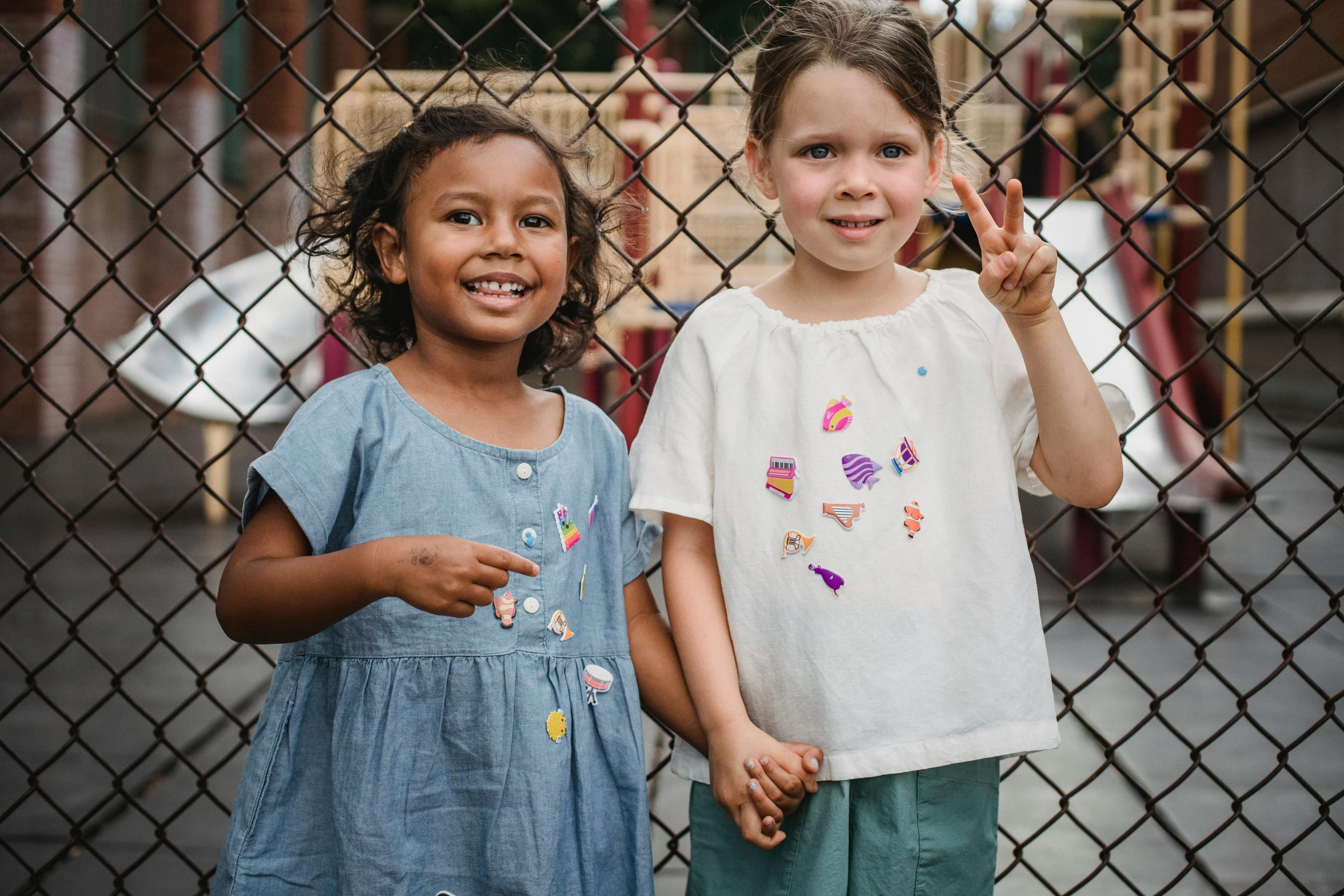 two little girls stand near a chain link fence, both holding their hands up