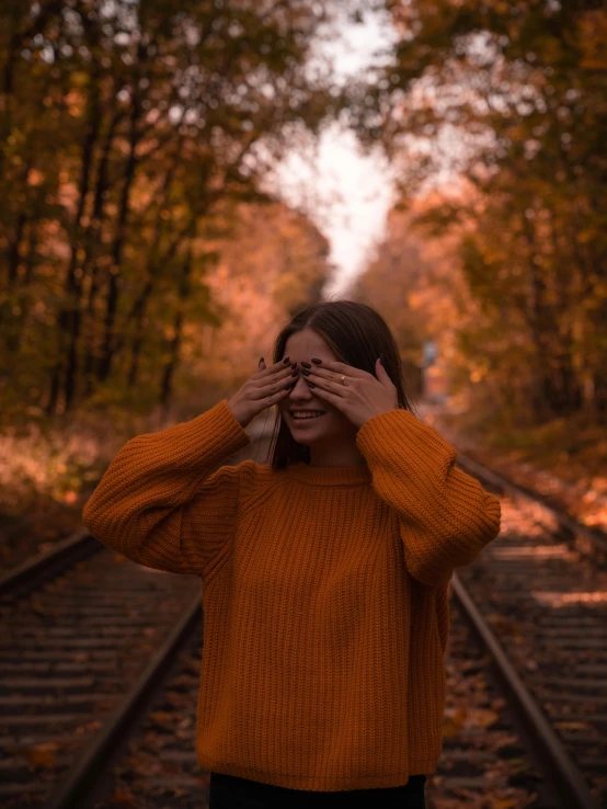 a woman covers her eyes as she walks along train tracks in autumn