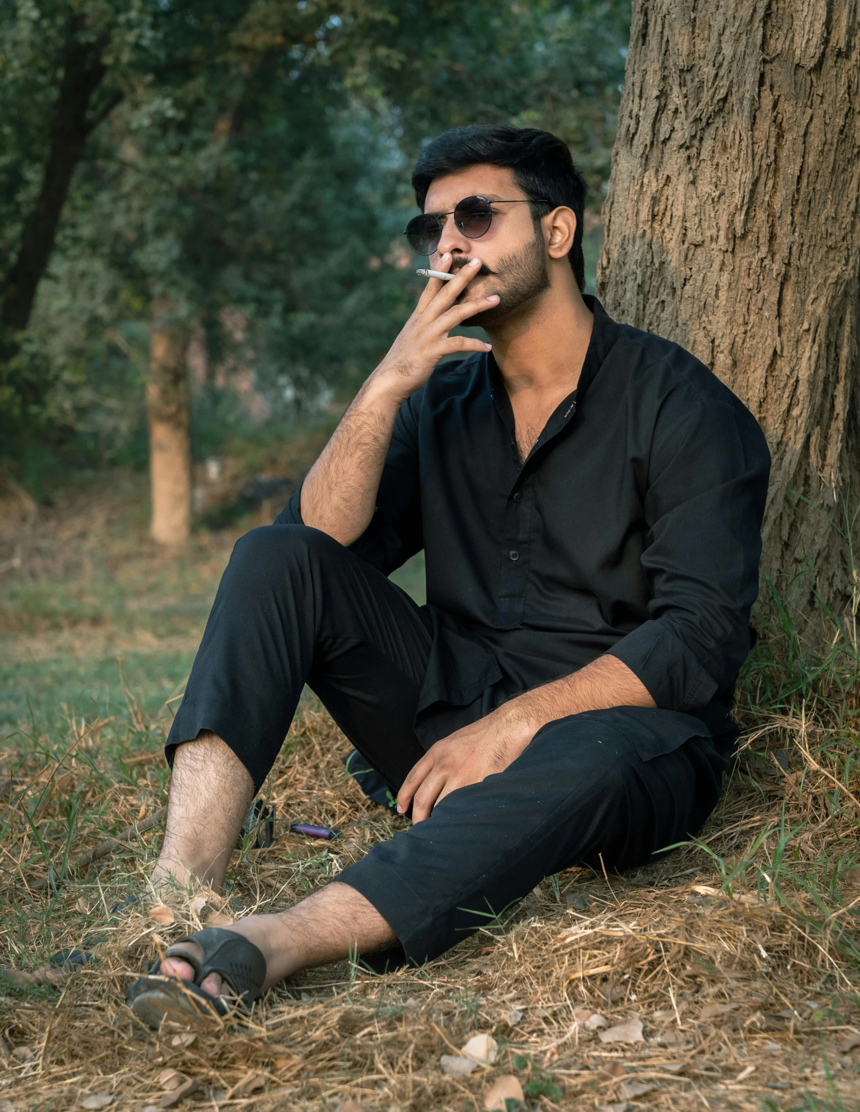 a man sits underneath a tree on the ground with his hand to his face while smoking a cigarette