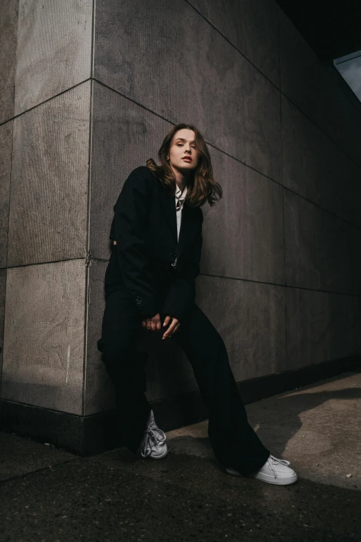 a woman sitting against a wall while wearing a black suit