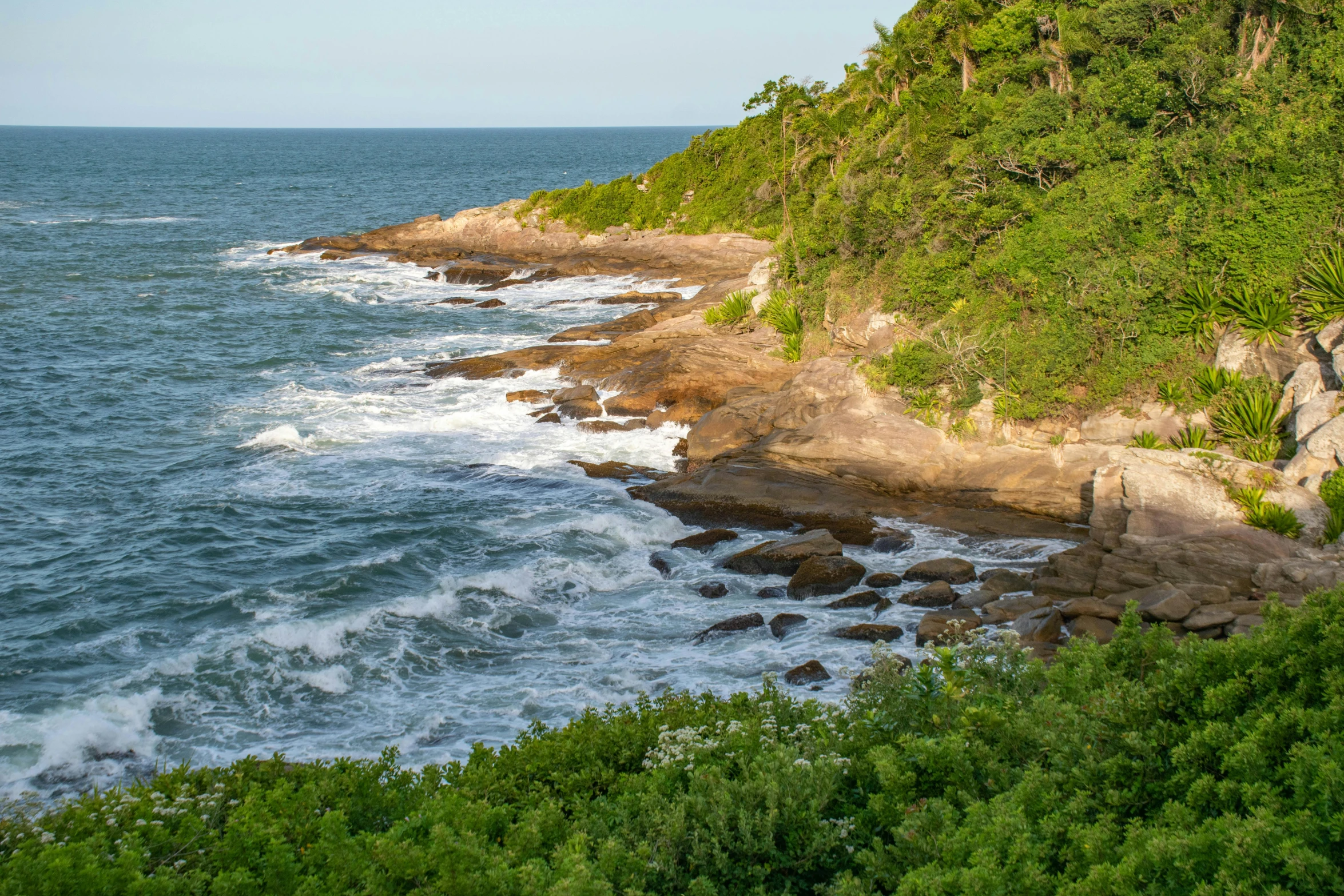 the view from the top of a rocky cliff, where some water is crashing against the shore and trees