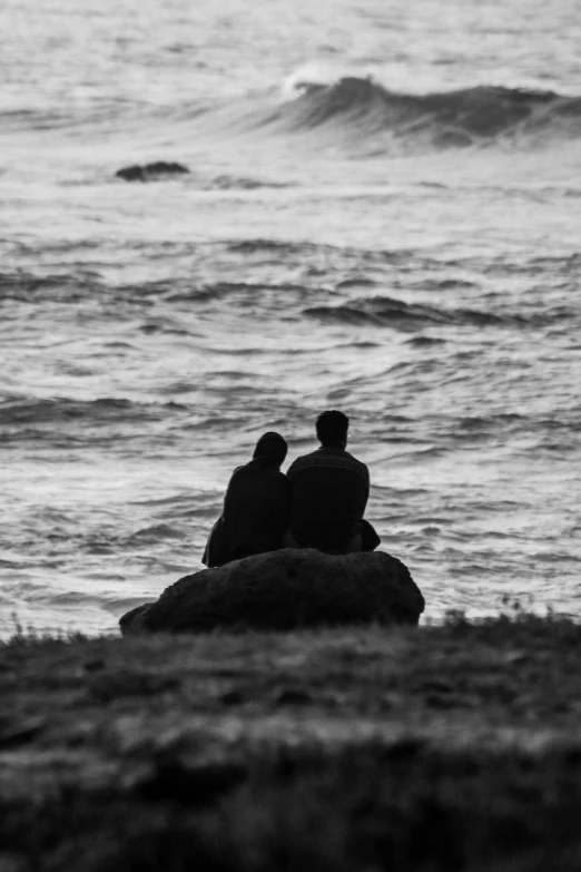 two people sitting on a rock by the ocean