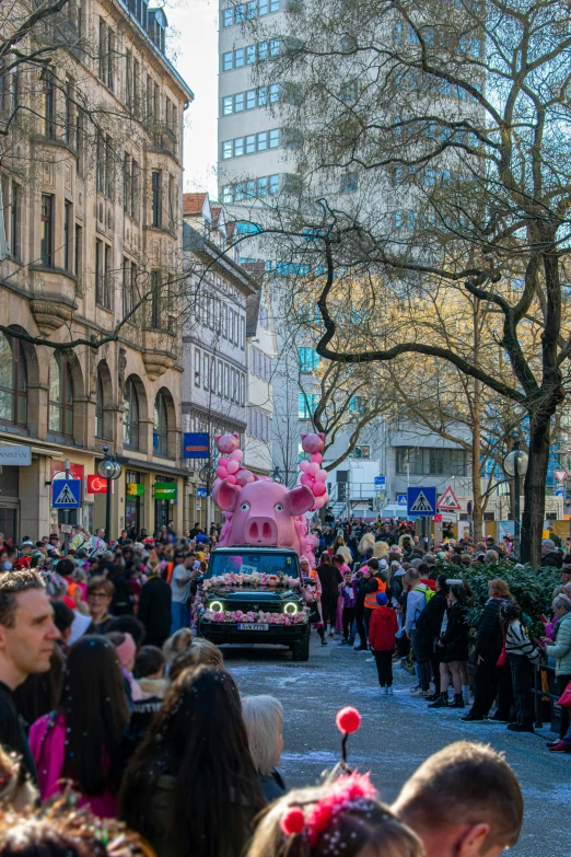 a street parade of people and vehicles with a pink horse in the middle of it