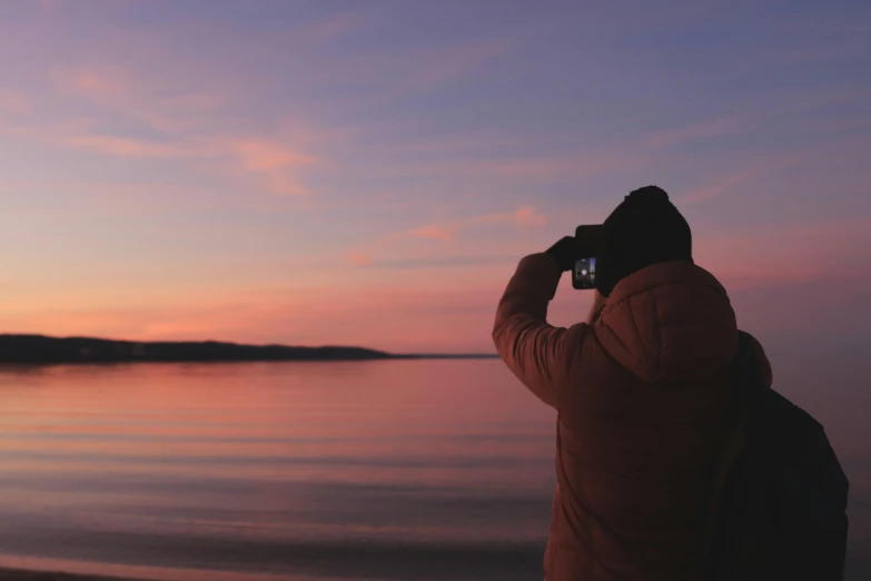 a man in winter jacket and hood taking pictures on the beach