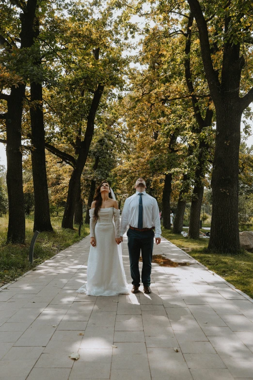 a bride and groom holding hands walking in the shade of trees