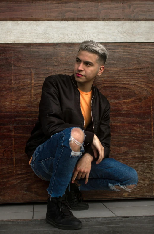 a young man sitting on a wooden crate wearing jeans and a black jacket