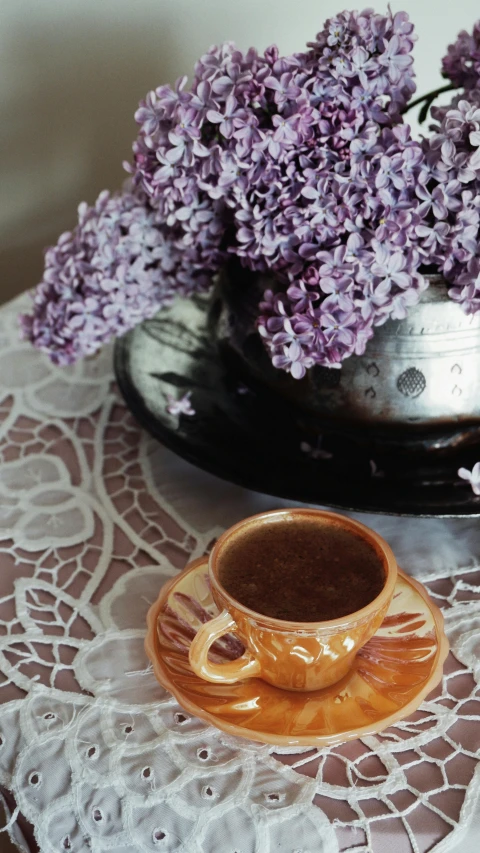 lilacs in an antique vase sit on a lace table cloth next to a coffee cup