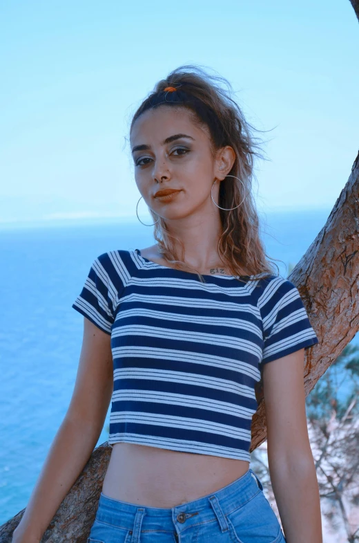 a young woman wearing a striped shirt near a tree with the ocean in the background