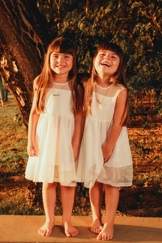 two girls wearing white dresses stand against a tree