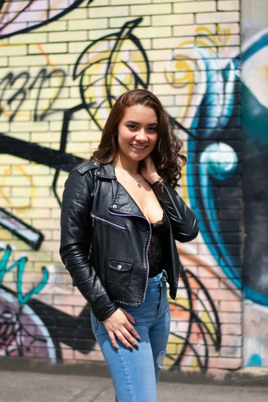 woman in jeans and black leather jacket posing near wall with graffiti