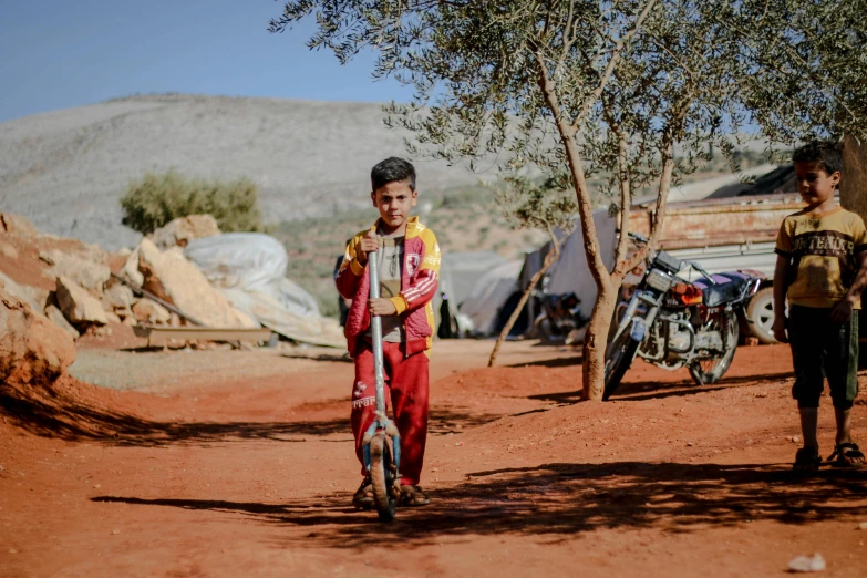 a boy holding a bat while standing next to another boy on a dirt road